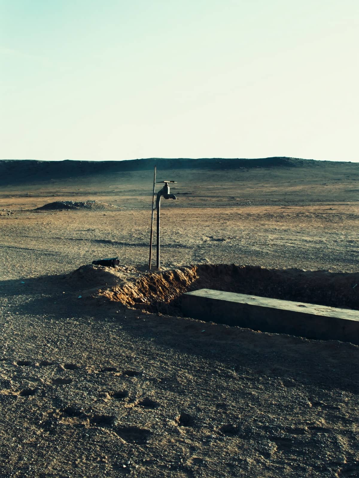 A lone standpipe and concrete watering trough on an empty Gobi gravel plain at dusk, the ground around them churned and dry